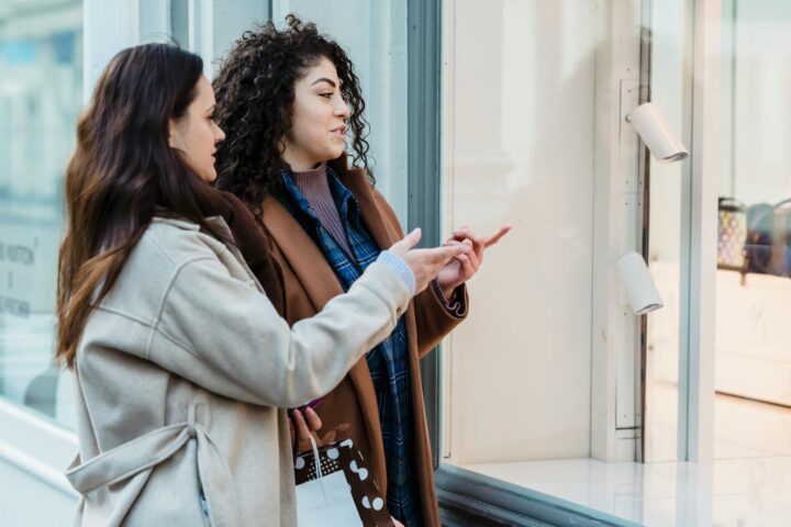 stylish young diverse female shopaholics pointing at store window on street